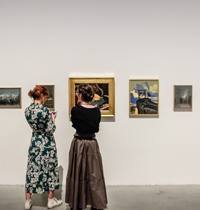 Zdjęcie. Dwie kobiety stoją przed obrazami Aleksandry Waliszewskiej. Ubrane a w długie sukienki. Rozmawiają ze sobą.s