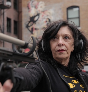 Starsza kobieta, brunetka celuje w kogoś bronią stojąc na podwórku murowanej kamienicy. Ubrana jest w czarną bluzę i bluzkę ze wzorem żółtych oczu. 