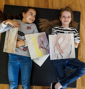 Zdjęcie. Dwie dziewczynki leżą na podłodze. Uśmiechają się. W ręku trzymają rysunki.