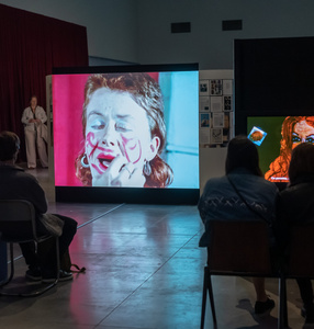 Zdjęcie. Kolorowe. Przyciemniona przestrzeń wystawy. Osoby siedzą tyłem do obiektywu i oglądają wideo na którym widać głowę kobiety, która maluje sobie twarz czerwoną szminką.