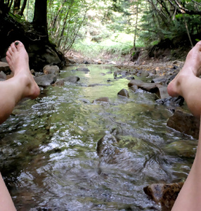 Zdjęcie. Dwie kobiece nogi od kolan po stopy, widziane po bokach zdjęcia. Między nogami widzimy rzekę. 