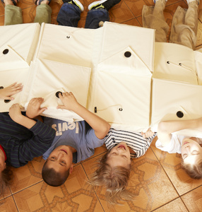 Zdjęcie. Czwórka dzieci leży na podłodze przykryte rozwiniętym pudełkiem z materiału w kolorze kremowym.