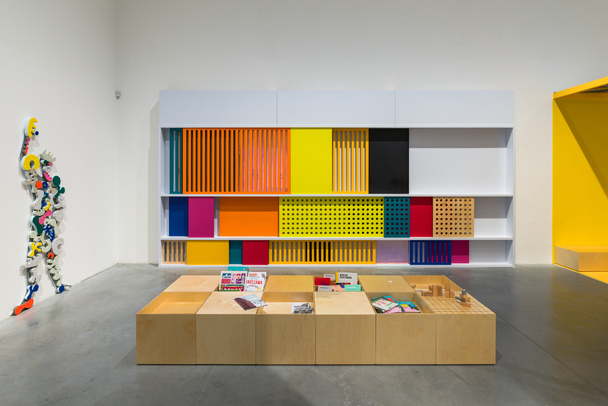 Zdjęcie przedstawiające półki z wielobarwnymi tablicami, na pierwszym planie pudła z drewna z książkami, po lewej na ścianie postać modulora z przyczepionymi kolorowymi gąbkami.