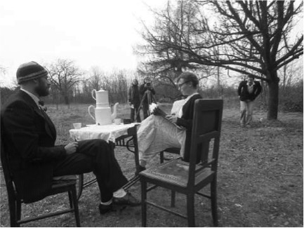 [czarno-białe zdjęcie przestawia kobietę i mężczyznę siedzących przy stole w przestrzeni ogrodu. W prawym górnym rogu zdjęcia widać duże drzewo, pod którym stoi męska postać]
