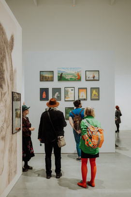 [zdjęcie 3 osób patrzących na prace wiszące na białej ścianie muzeum]
