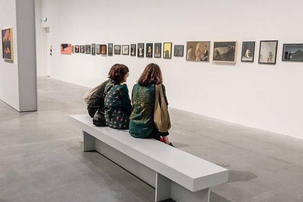 Zdjęcie. Dwie osoby zwiedzają wystawę Aleksandry Waliszewskiej. Siedzą na ławce przed obrazami zawieszonymi na ścianie.