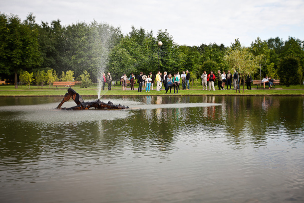 Ludzi zgromadzeni nad brzegiem jeziorka w Parku Rzeźby na Bródnie. Po środku jeziorka widać rzeźbę-fontannę, która przedstawia leżącą kobietę.