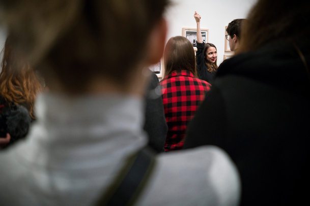Zdjęcie. Kobieta stoi wśród grupy osób: wyciąga rękę do góry, która widnieje nad tłumem.W tle widać obraz zawieszony na ścianie.