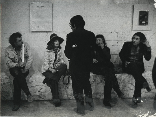 Photo. Black and white. Four people are sitting on a wall leaning against a white brick wall with artwork hanging on it.