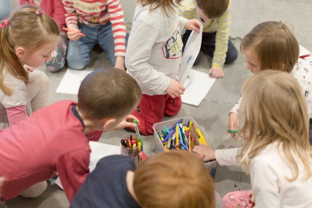 Zdjęcie. Grupa dzieci siedzi na podłodze i maluje coś na kartkach papieru.