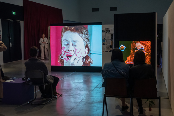 Photo. Color. A darkened exhibition space. People sit with their backs to the lens and watch a video showing a woman\'s head painting her face with red lipstick.