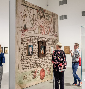 Zdjęcie. Ludzie zwiedzają wystawę Aleksandy Waliszewskiej. Dwie kobiety przyglądają się ścianie pełnej różnych obrazków i zapisków. Jakiś mężczyzna robi zdjęcie telefonem.