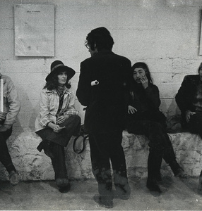 Photo. Black and white. Four people are sitting on a wall leaning against a white brick wall with artwork hanging on it.