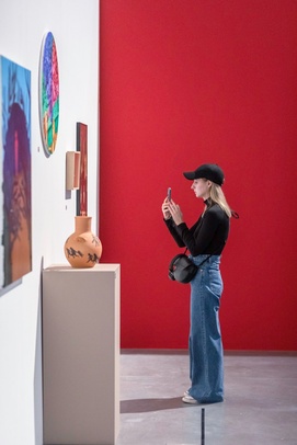 na zdjęciu widzka robi fotografię pracy telefonem, w tle czerwona ściana z wiszącymi na niej obrazami