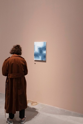 [na zdjęciu z wystawy osoba w ciemnobrązowym płaszczu patrzy na obraz zawieszony na ścianie w jasnobrązowym kolorze]