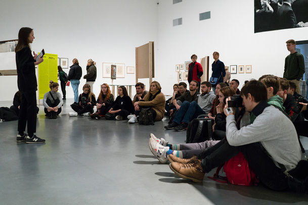 Zdjęcie. Edukatorka stoi przed grupą osób siedzących na ziemi w przestrzeni wystawowej Muzeum.
