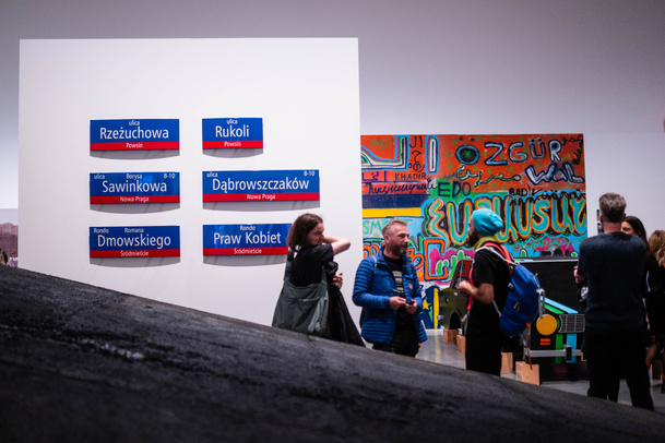 Zdjęcie. Trzy osoby rozmawiają na wystawie. Za nimi po lewej stronie biała ściana, na której znajduje się 6 tabliczek z nazwami warszawskich ulic. Za nimi po prawej stronie kolorowy obraz w stylu grafitti. 