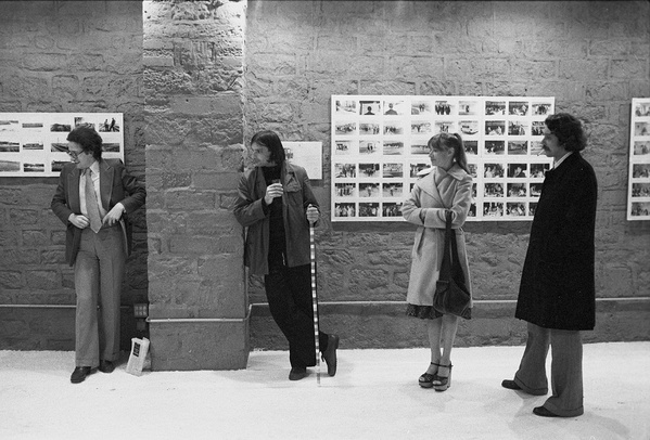 Zdjęcie. Czarno-białe. Cztery osoby na wystawie, przed ceglaną ścianą. Na ścianie małe zdjęcia oprawione grupowo w duże, białe ramy.