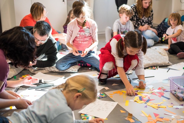 Zdjęcie. Dzieci wraz z rodzicami wyklejają prace z kolorowego papieru. Wszyscy siedzą bądź leżą na podłodze.