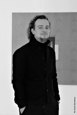 Zdjęcie czarno-białe, portret. Tomek Grabowski stoi na tle jasnej ściany. Jest ubrany na czarno. Uśmiecha się lekko.